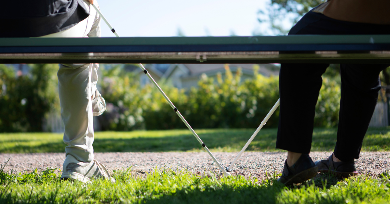 2 ihmistä istuu penkillä takaapäin kuvattuna, molemmilla on kädessään valkoiset kepit.